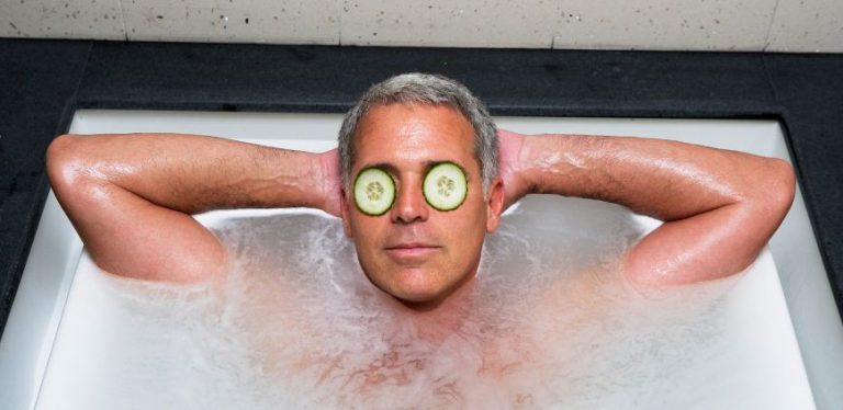 A man laying in a bath tub with his hands behind his head, and cucumber slices covering his eyes.