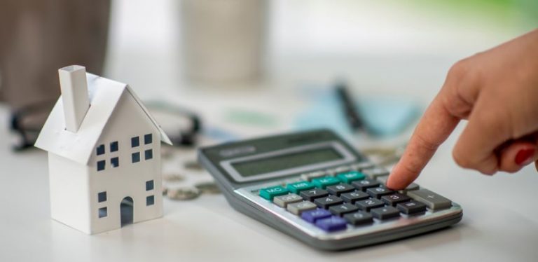 A small model of a house sitting on a table beside a calculator, someone's finger is pressing buttons on the calculator.