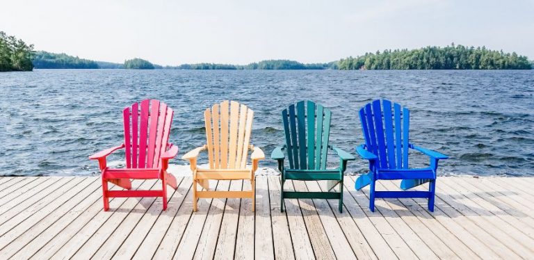 Four Adirondack chairs on a dock in front of a lake.