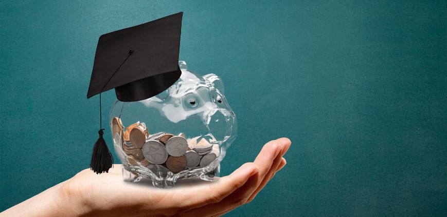 A clear piggy bank filled with coins, wearing a graduation cap being held in a person's hand.