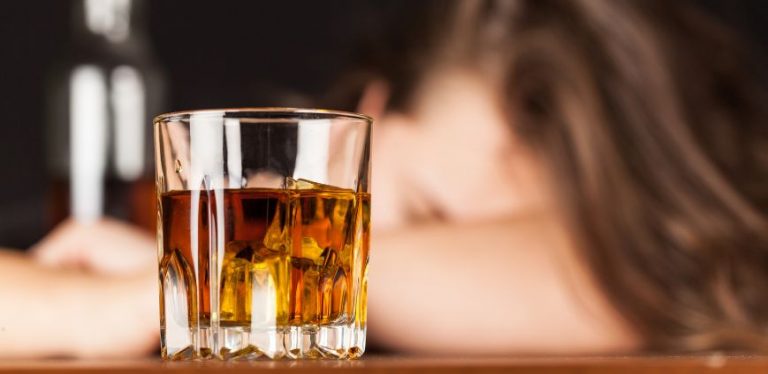 A glass of alcohol with a woman in the background. She has her head down on a table.