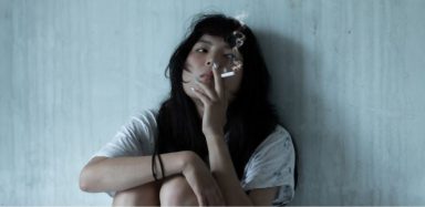 A woman sitting against a wall smoking a cigarette.