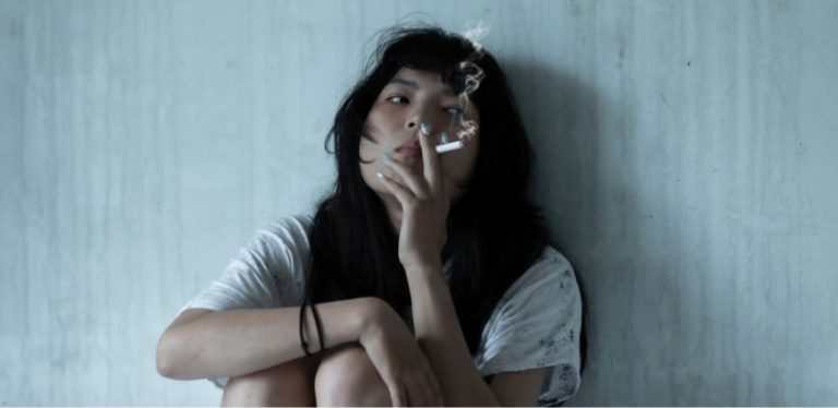 A woman sitting against a wall smoking a cigarette.