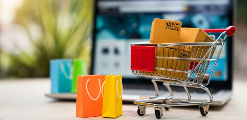 A miniature shopping cart full of bags and boxes, with a laptop in the background.
