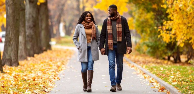 A couple holding hands and walking down a sidewalk in the Fall. The foliage is very colorful in shades of orange and yellow.