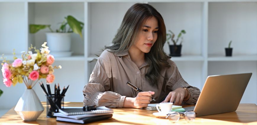 A woman sitting at a desk, reading off a laptop and taking notes.